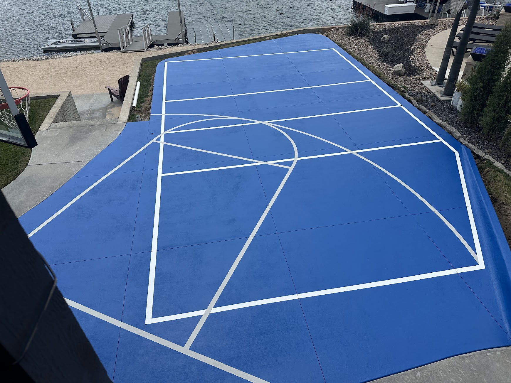 Custom outdoor basketball and sport court featuring a bright blue surface, white court markings, and a lakeside setting.